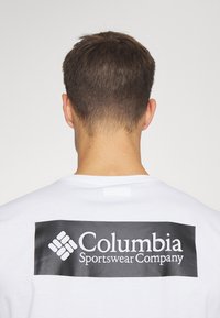 Hvit bomull t-skjorte med et stort svart rektangulært logo på baksiden med teksten "Columbia Sportswear Company" i hvitt.