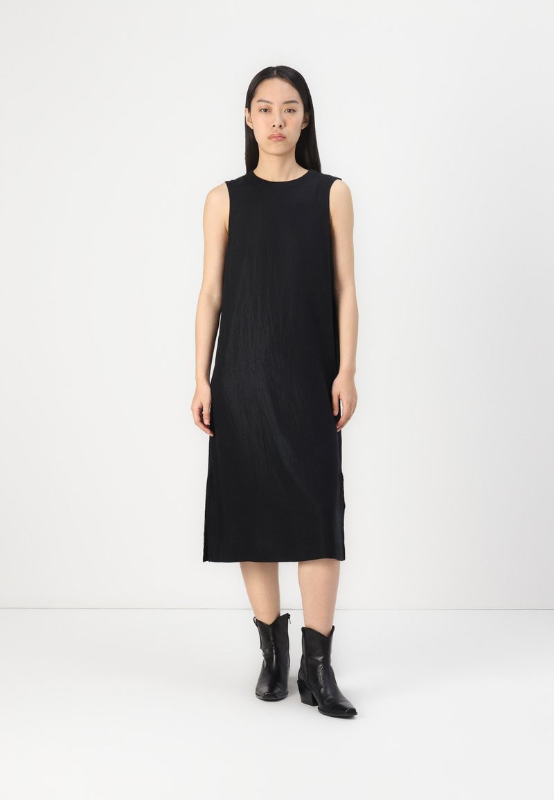 Schwarzes ärmelloses Kleid mit geradem Schnitt, aus strukturiertem Stoff. Getragen mit schwarzen Western-Stiefeletten, vor einem klaren Hintergrund.