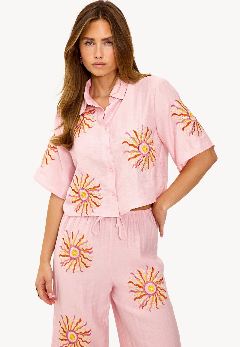 Ensemble en lin rose comprenant une chemise à boutons courte et un pantalon ample, décoré de broderies solaires vibrantes en jaune, orange et rose.