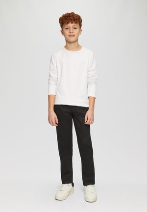 Witte lange mouwen shirt, zwarte broek en witte sneakers gedragen door een jongen met krullend rood haar die tegen een effen achtergrond staat.