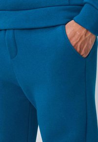 Pantalon de survêtement turquoise avec une texture douce, doté d'une poche avant et d'une ceinture à cordon. Lignes épurées et détails de design minimalistes.