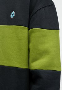 Artimas juodos ir žalios sportinės maikutės rankovės vaizdas su mažu mėlynu siuvinėtu liepsnos logotipu viršutinėje juodoje dalyje.