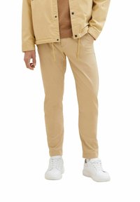 Beige bomullsbukser med en avsmalnande passform, snörningsdetaljer och sidofickor, tillsammans med vita sneakers och en brun tröja under en beige jacka.