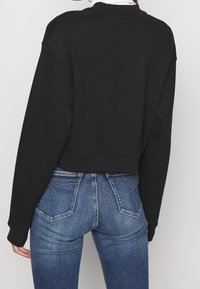 Person klädd i en svart långärmad kort sweatshirt och hög midja blå denimjeans, sedd bakifrån.
