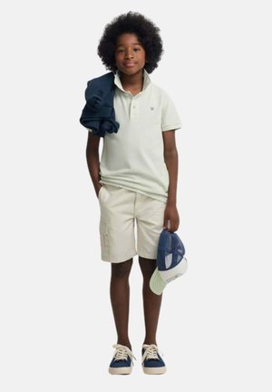 Niño con afro llevando polo claro, pantalones cortos beige, zapatillas azules, sosteniendo gorra azul y blanca, sudadera azul marino sobre el hombro.