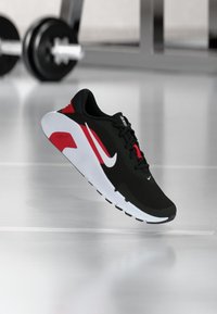 Μαύρο και κόκκινο αθλητικό παπούτσι Nike για τρέξιμο με λευκή σόλα να αιωρείται πάνω από λαμπερό δάπεδο, με βάρη γυμναστικής θολά στο φόντο.