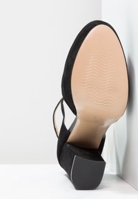 Černá semišová bota na vysokém podpatku s kulatou špičkou a otevřenou zadní částí. Obsahuje hladkou béžovou podrážku a široký podpatek.