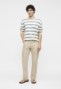 Jeune homme portant une chemise blanche et grise à rayures, manches longues, un pantalon beige et des sandales marron clair, debout avec une main dans la poche.