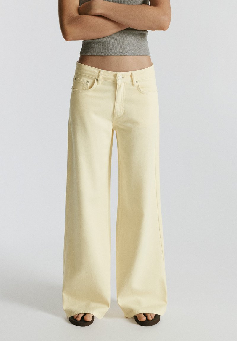 Personne portant un pantalon large jaune clair, un top court gris et des sandales noires, debout, les bras croisés sur un fond uni.