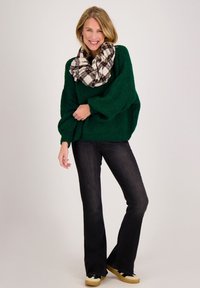 Vrouw die lacht, een groene oversized trui, zwarte uitlopende spijkerbroek, geruite sjaal en sneakers dragend, staand tegen een effen achtergrond.