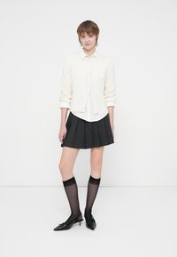 Cardigã branco sobre uma camisa branca, saia plissada cinza, meias altas transparentes pretas até ao joelho e sapatos pretos de bico fino com detalhes em laço.