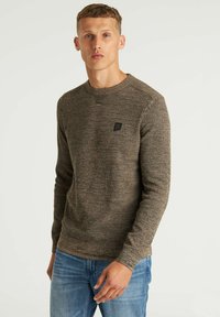 Brauner gestrickter Pullover mit einem runden Ausschnitt, strukturiertem Stoff und einem kleinen schwarzen Logo-Patch auf der Brust, kombiniert mit hellblauen Jeans.