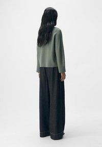 Vrouw met lang donker haar, gekleed in een groene trui en een wijdvallende zwarte broek met streepjes, staand met de rug naar de kijker op een effen achtergrond.