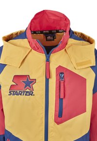 Kolorowa kurtka z żółtymi, czerwonymi i niebieskimi panelami, zapinana na zamek z przodu, z logo na piersi; posiada odpinany kaptur i kieszeń.