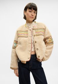Kurtka w kolorze kremowym, wykonana z teksturowanego materiału przypominającego polar. Posiada wielokolorowe poziome paski oraz mosiężne napy. Krótki fason.