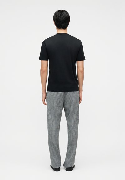 Άνδρας στέκεται με την πλάτη, φοράει μαύρο κοντομάνικο πουκάμισο και γκρι παντελόνι με ρίγες, μπροστά σε απλό λευκό φόντο.