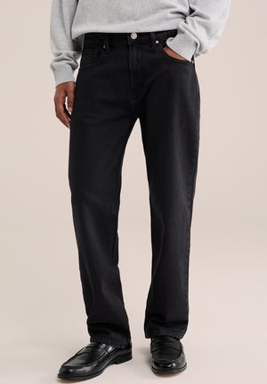 Homme portant un jean noir droit, des mocassins en cuir noirs et une chemise à manches longues gris clair, une main dans la poche.