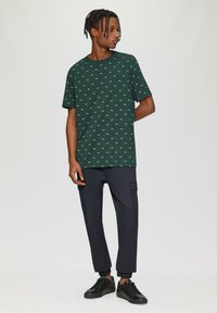 Groene katoenen t-shirt met een wit geometrisch patroon, ronde hals, en korte mouwen, gecombineerd met zwarte cargobroeken en zwarte sneakers.