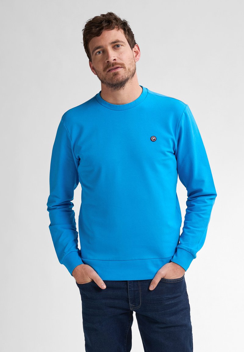 Felpa blu in morbido tessuto, con maniche lunghe, collo rotondo e un piccolo logo sul petto. Abbinata a jeans scuri.
