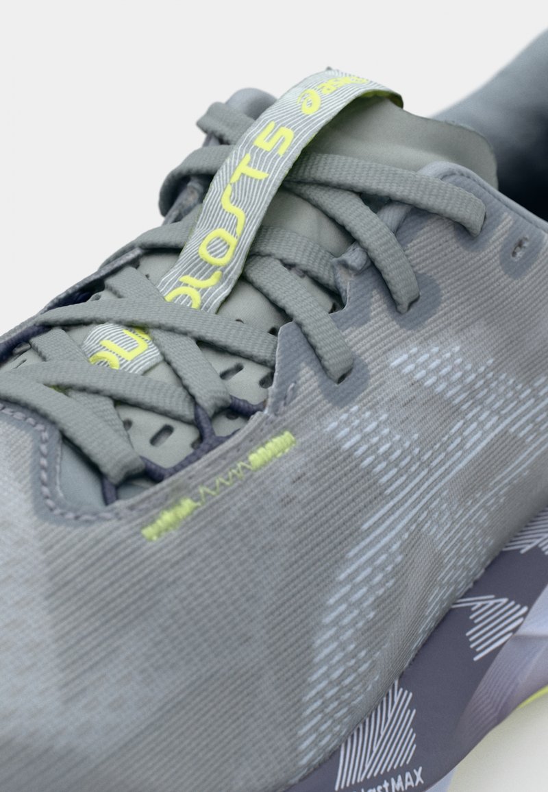 Grijze hardloopschoen met een bovenwerk van textuurstof, opvallende veters en neongele accenten. Beschikt over een paarse zool met geometrisch patroon.