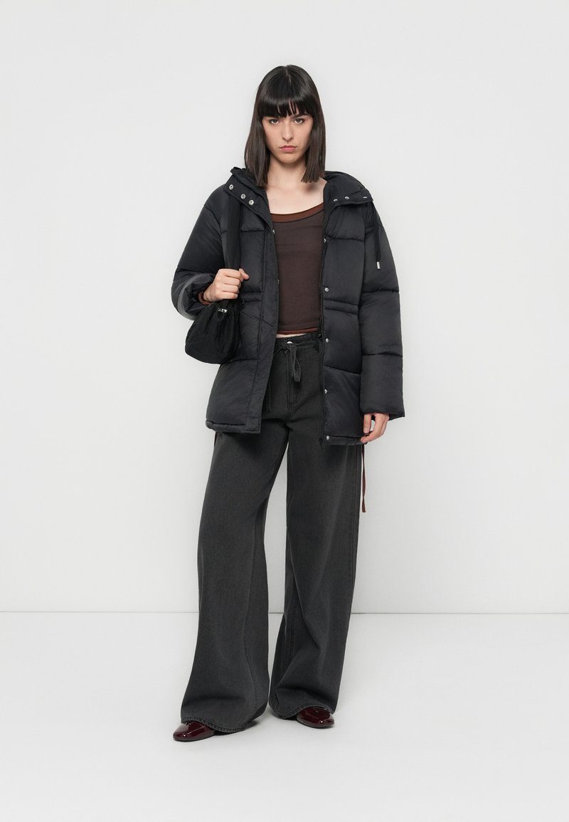 Schwarze Daunenjacke mit Kapuze, braunes tailliertes Oberteil und lockere graue weit geschnittene Hose. Die Accessoires umfassen eine schwarze Tasche und bordeauxfarbene Schuhe.