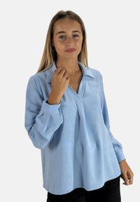 Blouse bleu clair texturé avec un col pointu et des manches longues légèrement bouffantes, dotée d'une coupe décontractée et d'un décolleté en V.