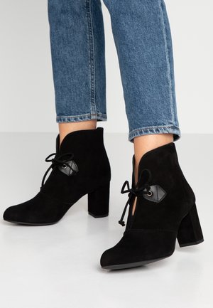 Personne portant un jean bleu et des bottines en daim noir à lacets avec talons blocs, debout sur un sol blanc.