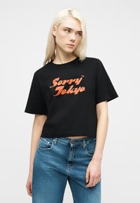 Νέα γυναίκα με ξανθά μαλλιά, που φοράει μαύρο κοντό μπλουζάκι με πορτοκαλί κείμενο "Sorry Tokyo" και μπλε τζιν, ποζάροντας μπροστά σε λευκό φόντο.