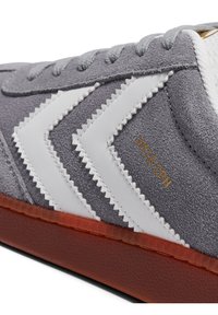 Pantof sport din piele întoarsă gri cu dungi albe în zigzag și text auriu. Prezintă o talpă din cauciuc portocaliu texturată și șireturi gri.