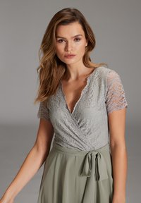 Top in pizzo grigio con maniche corte, scollo a V e design avvolgente; abbinato a una gonna verde fluida con dettaglio in vita annodato.