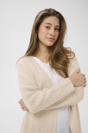 Cardigan en tricot beige avec un motif en chevron, ouverture sur le devant et poignets côtelés. Superposé à un t-shirt blanc, texture douce visible.
