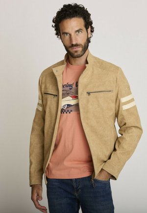 Homme aux cheveux bouclés et foncés avec une barbe, portant une veste beige zippée sur un t-shirt graphique pêche et un jean bleu, debout avec une main dans la poche.