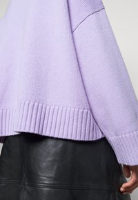 Pull en tricot lavande avec ourlet et poignets côtelés porté sur une jupe en cuir noir, main reposant à côté de la jupe.