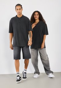 To modeller har på seg oversized svarte polo skjorter. Den ene har mørke denimshorts og svarte sneakers; den andre har lyse grå vidde bukser og hvite sneakers.