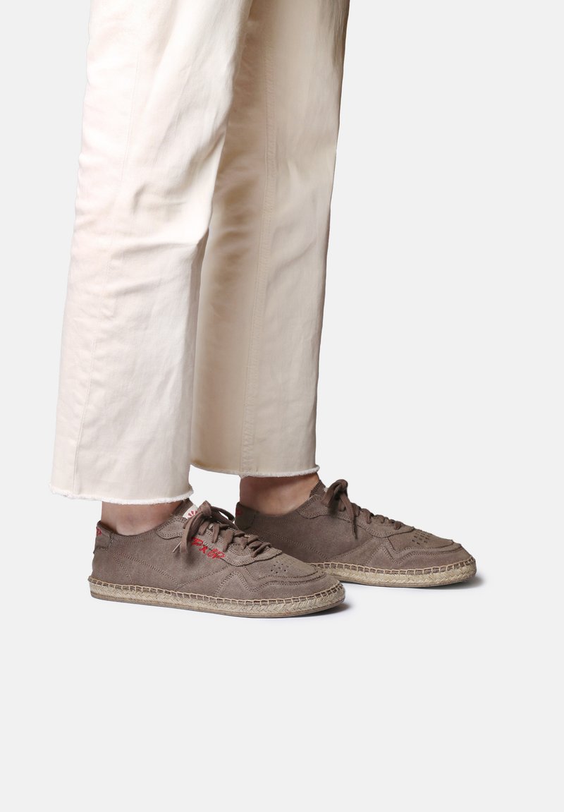 Personne portant un pantalon beige raccourci et des baskets en daim marron avec des semelles enveloppées de jute sur un fond uni.