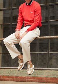 Rotes Strick-Polo mit langen Ärmeln und schwarzem Kragen, helle beige Hose und beige/violette Sneaker mit weißen Akzenten, sitzend auf einem Geländer.