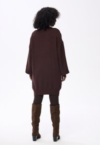 Vrouw met krullend haar, gekleed in een losse donkerbruine sweaterjurk, donkere panty's en kniehoge bruine suède laarzen, staande met haar rug naar de camera.