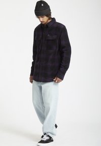 Camisa de franela a cuadros negros y morados con dos bolsillos en el pecho, combinada con jeans rectos de un azul claro y zapatillas negras. Se lleva un gorro negro.