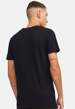 Man met kortgeknipte haren die een effen zwarte korte mouw T-shirt draagt, met zijn rug naar voren, waarbij tatoeages op zijn linkerarm zichtbaar zijn tegen een witte achtergrond.