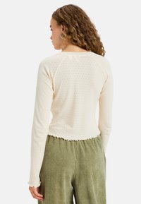 Vrouw met krullend haar die een crèmekleurige lange mouw top met een klein oogje patroon en olijfgroene wijde corduroy broek draagt.