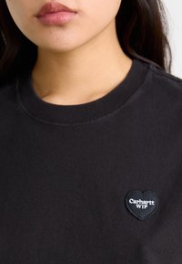 Svart bomull t-skjorte med rund halslinning og korte ermer, med et hjerteformet svart merke med hvit tekst "Carhartt WIP" på brystet.