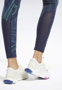 Leggings blu scuro aderenti con motivi geometrici verdi e blu, abbinati a sneaker bianche con tomaia texturizzata e suole colorate.