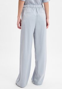 Persoon in lichtgrijze wijde broek met elastische tailleband, gezien van achteren tegen een effen witte achtergrond.