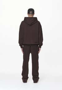 Bruine hoodie met capuchon en losse pasvorm, gecombineerd met bijpassende bruine joggingbroek met zijnaden. De achtergrond is effen wit.