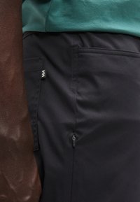 Σκούροι ναυτικοί παντελόνια με πλαϊνή τσέπη με φερμουάρ, κατασκευασμένα από υφασμάτινο ύφασμα, σε συνδυασμό με πράσινο βαμβακερό τοπ.