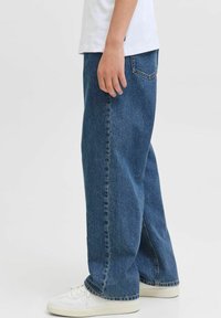 Jean bleu baggy en denim avec une coupe décontractée, comportant cinq poches et des détails de couture subtils. Associé à des baskets blanches.