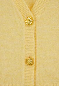 Dzeltens vilnas kardigans ar divām zelta saules formas pogām, V veida kakla izgriezumu, ribotu apkakli un mīkstu, teksturētu audumu.