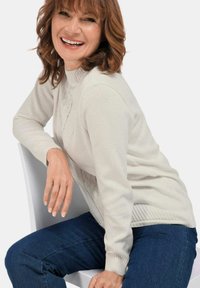 Hellebeiger Pullover mit strukturiertem Muster und geripptem Saum. Getragen mit dunkelblauen Jeans, hat lange Ärmel und einen hohen Halsausschnitt.