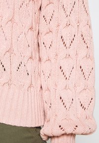 Pull en maille rose clair avec un motif texturé en losanges. Il comporte des poignets et un ourlet côtelés, mettant en valeur un matériau doux et respirant.