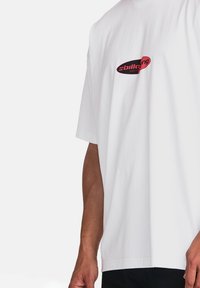 Hvid bomulds T-shirt med et sort og rødt logo på brystet. Korte ærmer, afslappet pasform og glat tekstur uden synlige mønstre.